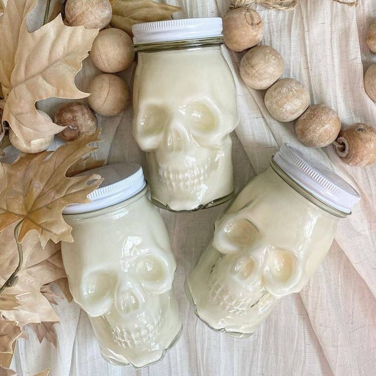 Three Skull Candle jars, crafted from glass with white lids and creamy soy wax, are set on light fabric among dried leaves and wooden beads—ideal as fall-inspired decor.