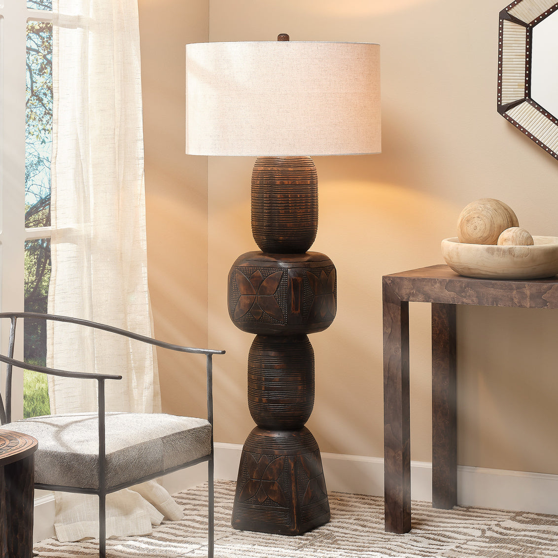 A tall, sculpted Totem Floor Lamp crafted from mango wood with a beige shade stands by a window, next to a wooden table adorned with decorative bowls and a modern chair. Sunlight filters through sheer curtains.
