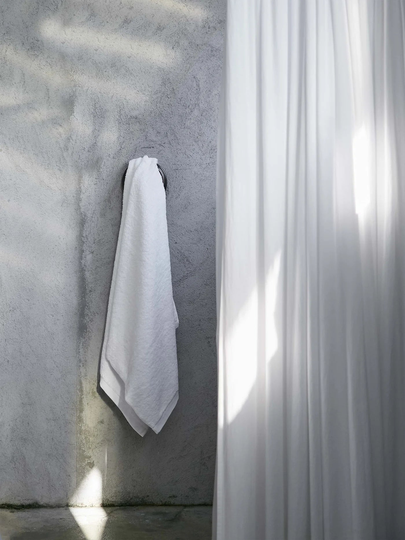 A smoke-colored Riviera Cotton Bath Towel, garment-dyed with non-toxic dyes, hangs on a hook against a textured gray wall as soft sunlight filters in. On the right, a white shower curtain gently billows into view.