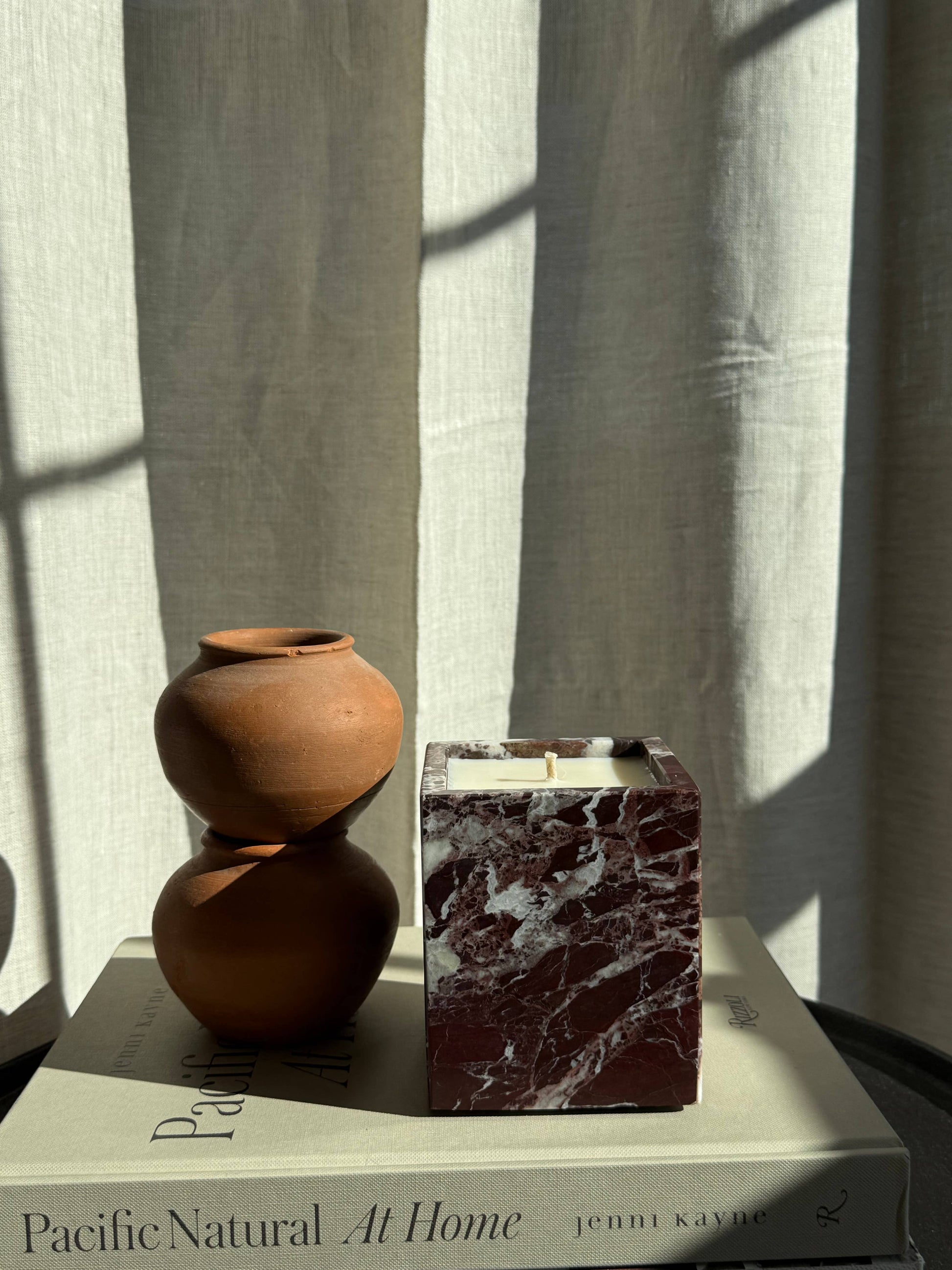 A small brown stacked clay vase and a Marble Candles holder rest on the beige book "Pacific Natural At Home" by Jenni Kayne, with sunlight and curtain shadows in the background.