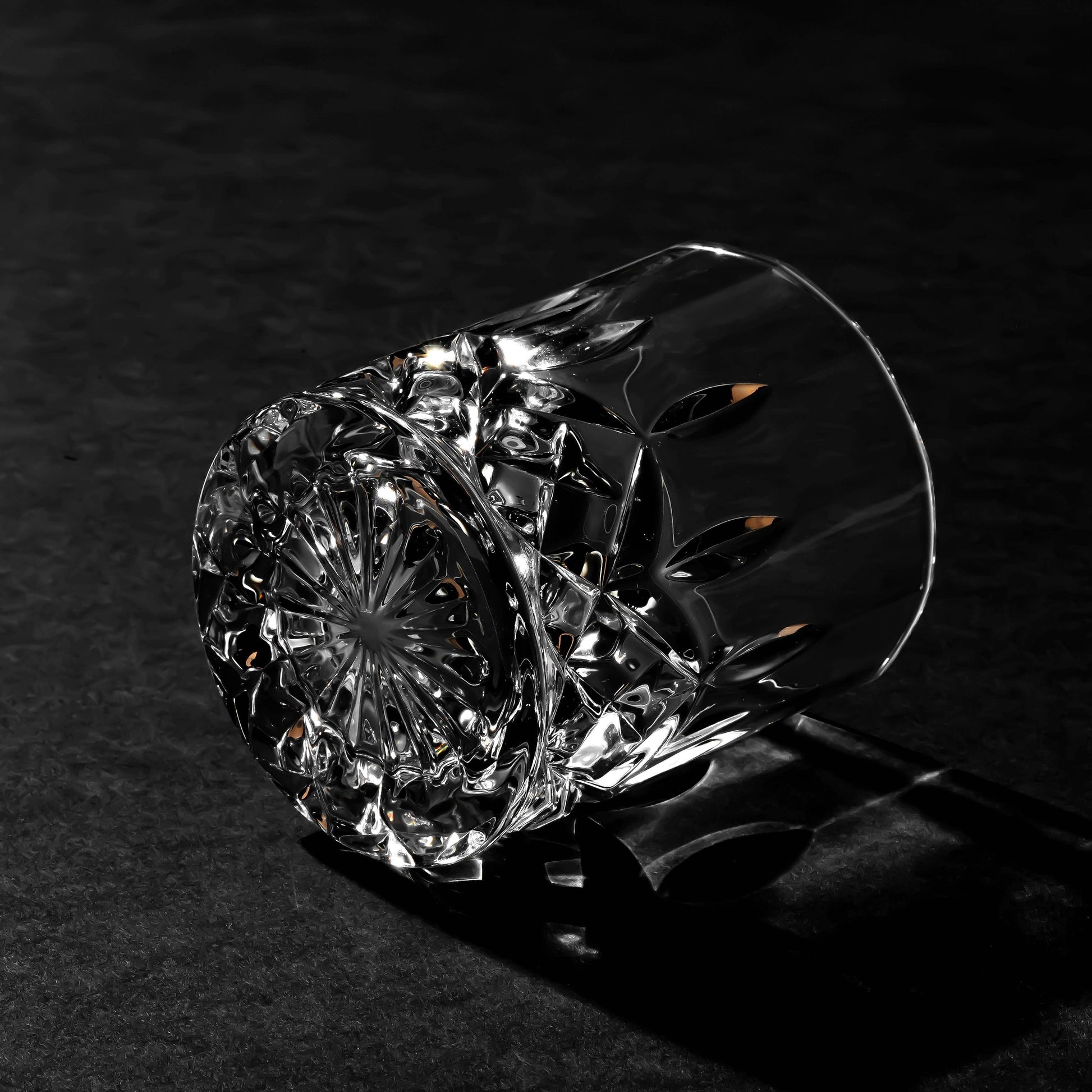 The Crystal Cut Old Fashioned Whiskey Glass 10oz (1pc) rests on its side atop a dark textured surface, with light highlighting its detailed cut patterns—an elegant gift for whiskey lovers.