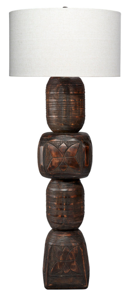 The Totem Floor Lamp features a carved base crafted from dark mango wood with geometric patterns and is topped with a large, white cylindrical lampshade.