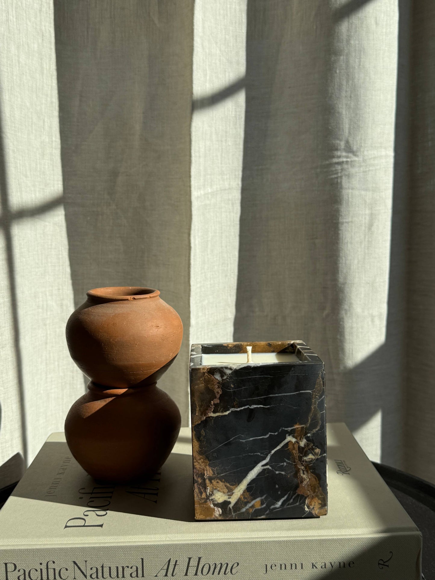 A small stack of two brown clay pots and a Marble Candles vessel rests atop the book "Pacific Natural At Home" by Jenni Kayne, set against beige curtains with sunlight streaming in.