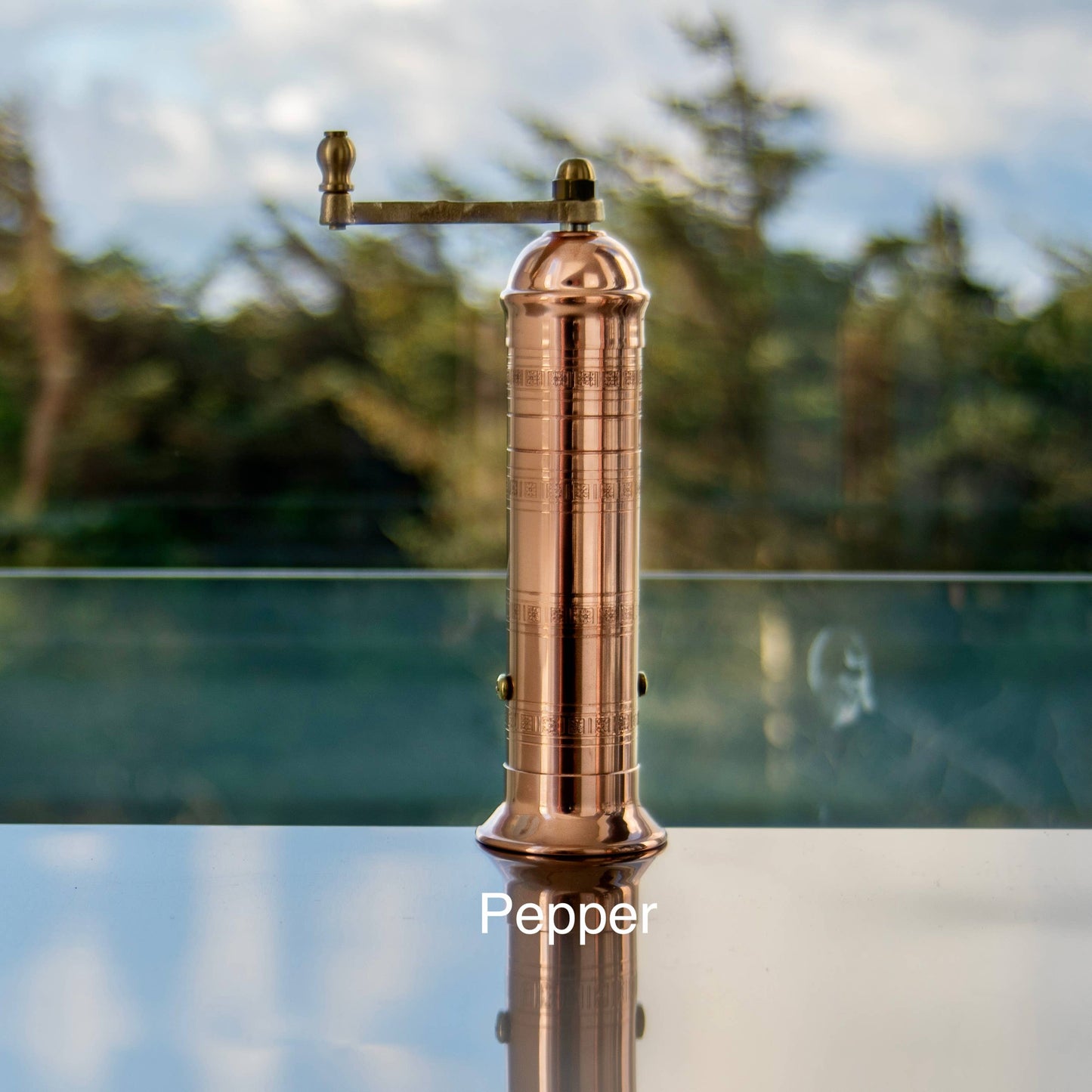 The original 'Copper Mill' - 9" pepper grinder with a hand crank sits labeled “Pepper” on a reflective surface, set against a blurred outdoor backdrop of greenery and sky.