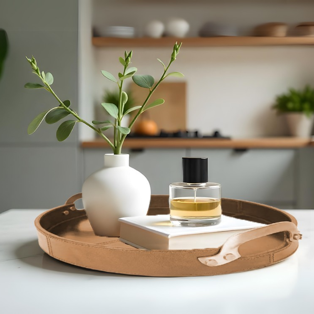 A small white vase with green stems, a perfume bottle, and a white book sit on the Round Leather Stitched Tray with Brass Handle atop a table. A modern kitchen with wooden shelves and plants is blurred in the background.