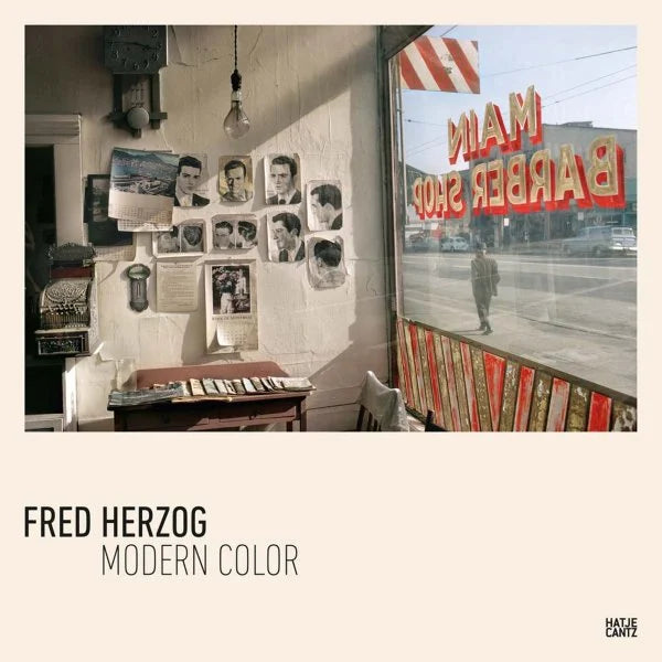 Experience the timeless charm of a vintage barbershop captured in Fred Herzog’s “Modern Color.” This striking 10.6″x10.6″ presentation features black-and-white portraits lining the walls, a well-worn chair, and a grand window bearing the name MAIN BARBER SHOP, with a man’s reflection gracing the glass. “Modern Color by Fred Herzog” beautifully blends nostalgic atmosphere with contemporary aesthetics.
