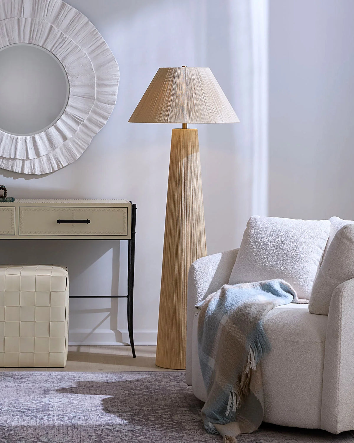 Modern living room with a tall Jute Floor Lamp featuring a 3-way socket for customizable lighting, a white round mirror, cream console table, white cushioned ottoman, textured white sofa, and a blue and beige throw blanket draped over the sofa.