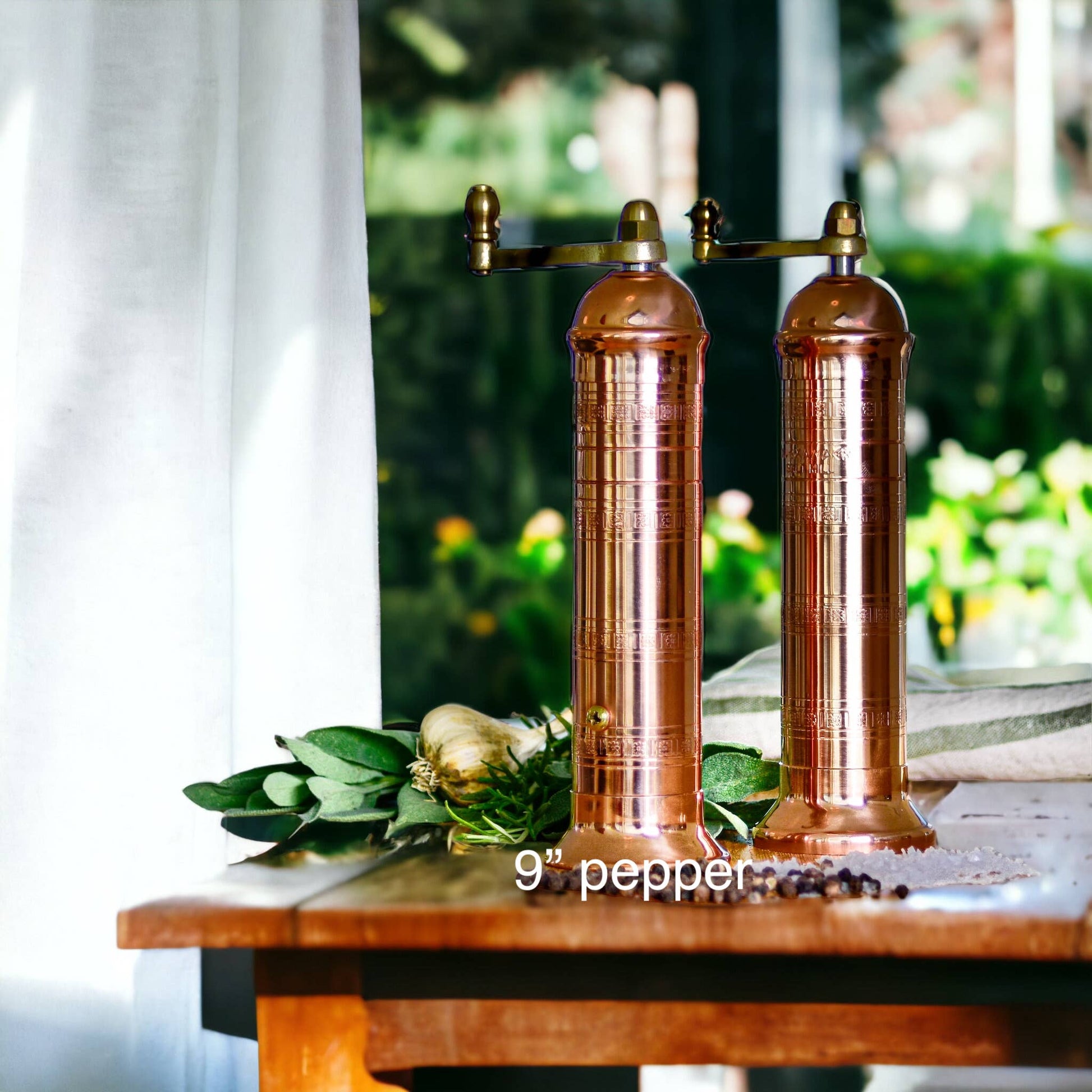 Two original 'Copper Mill' 9" pepper grinders from Alexander Handcrafted Mills sit on a wooden table by a sunlit window, accompanied by fresh herbs and garlic for a cozy, inviting atmosphere.
