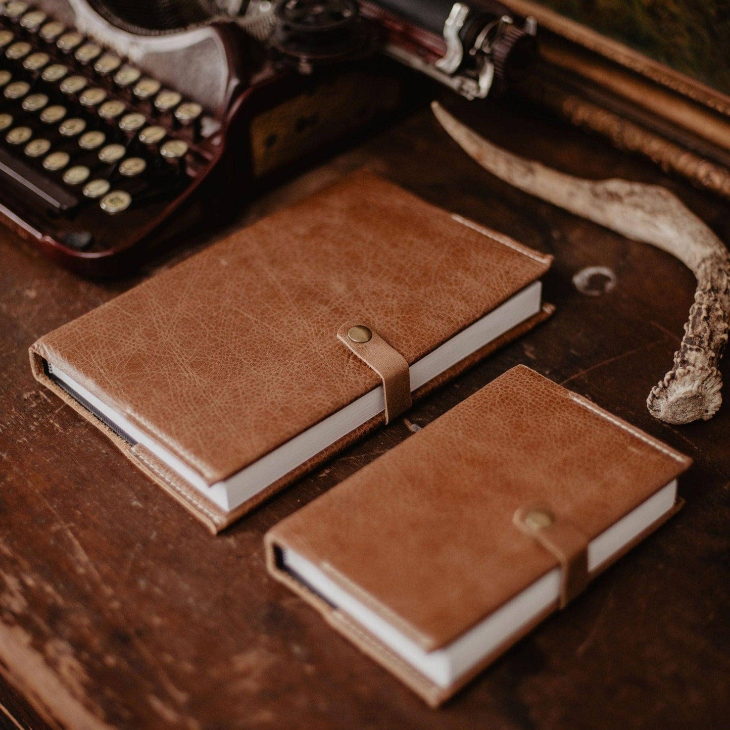 Two brown handcrafted Writer’s Journals—one large, one small—sit on a wooden desk beside an old typewriter and deer antler, each with a buttoned strap closure. A timeless custom branded keepsake for any desk.