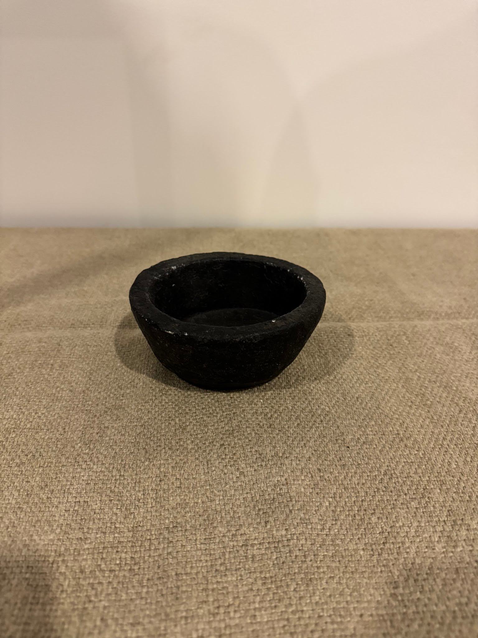The Black Bowl, small and round, rests on textured beige fabric with a plain light-colored wall in the background.
