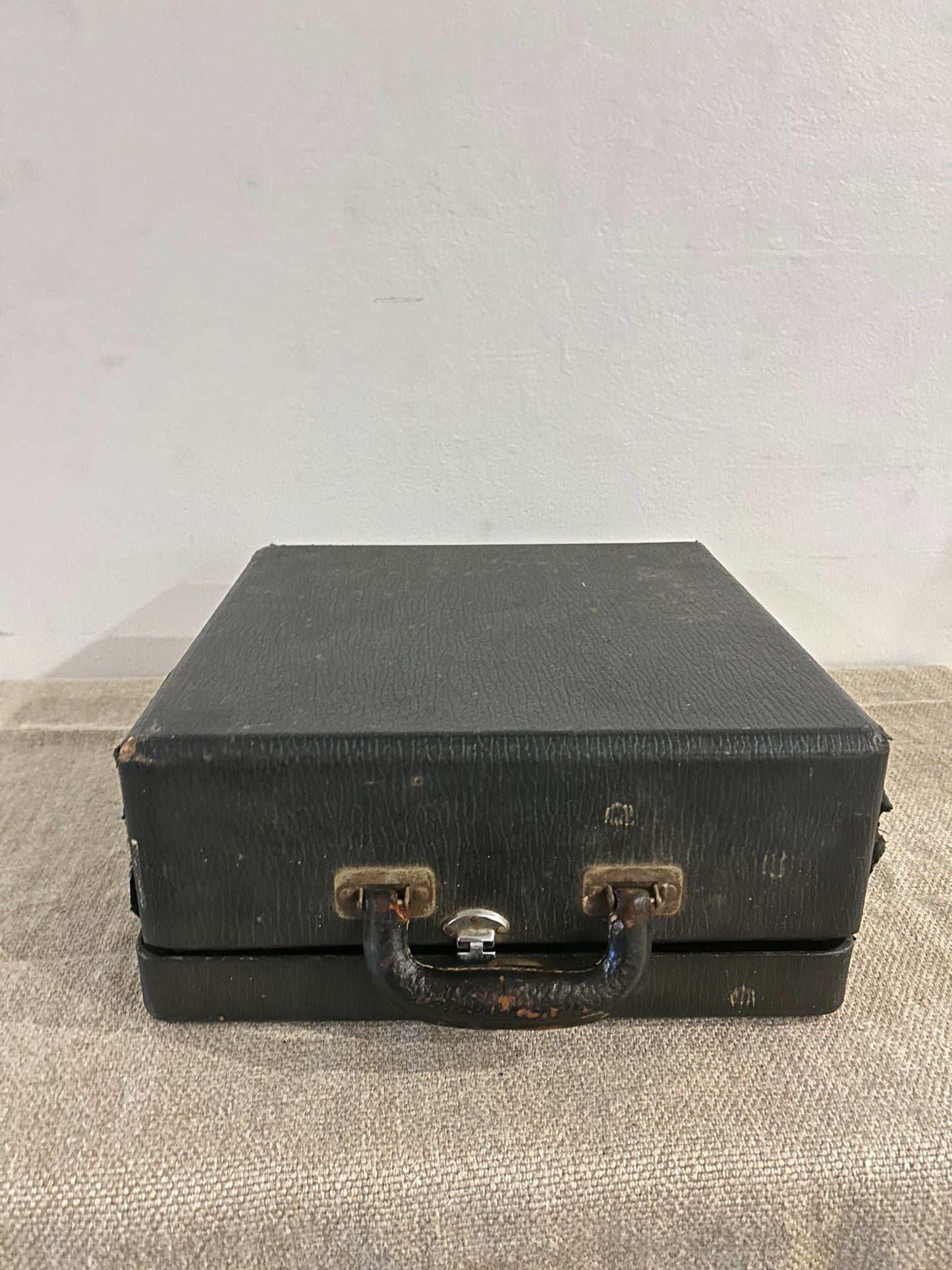 A small, square, black typewriter suitcase—once carrying the Black Typewriter—sits on beige fabric, its worn handle and metal clasps adding vintage charm against a plain white wall.