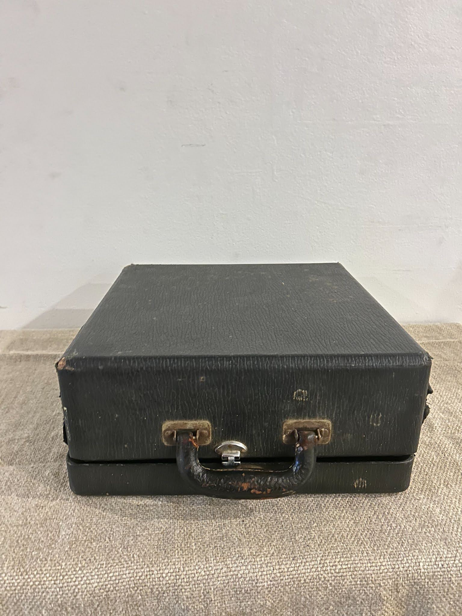 A small, square, black typewriter suitcase—once carrying the Black Typewriter—sits on beige fabric, its worn handle and metal clasps adding vintage charm against a plain white wall.