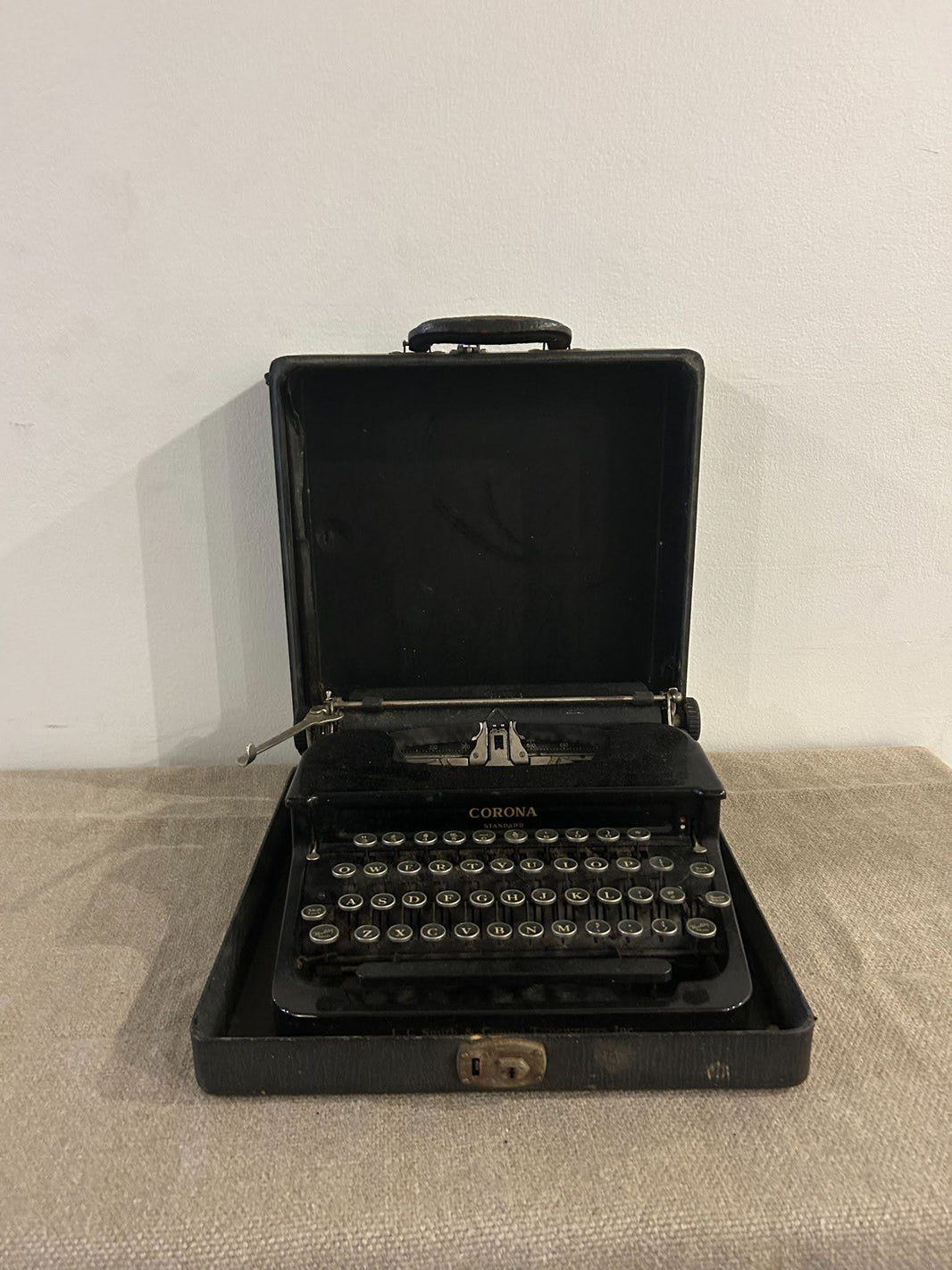 The Black Typewriter is a vintage portable model displayed open in its carrying case on a beige surface, with keys and handle visible against a plain white wall.
