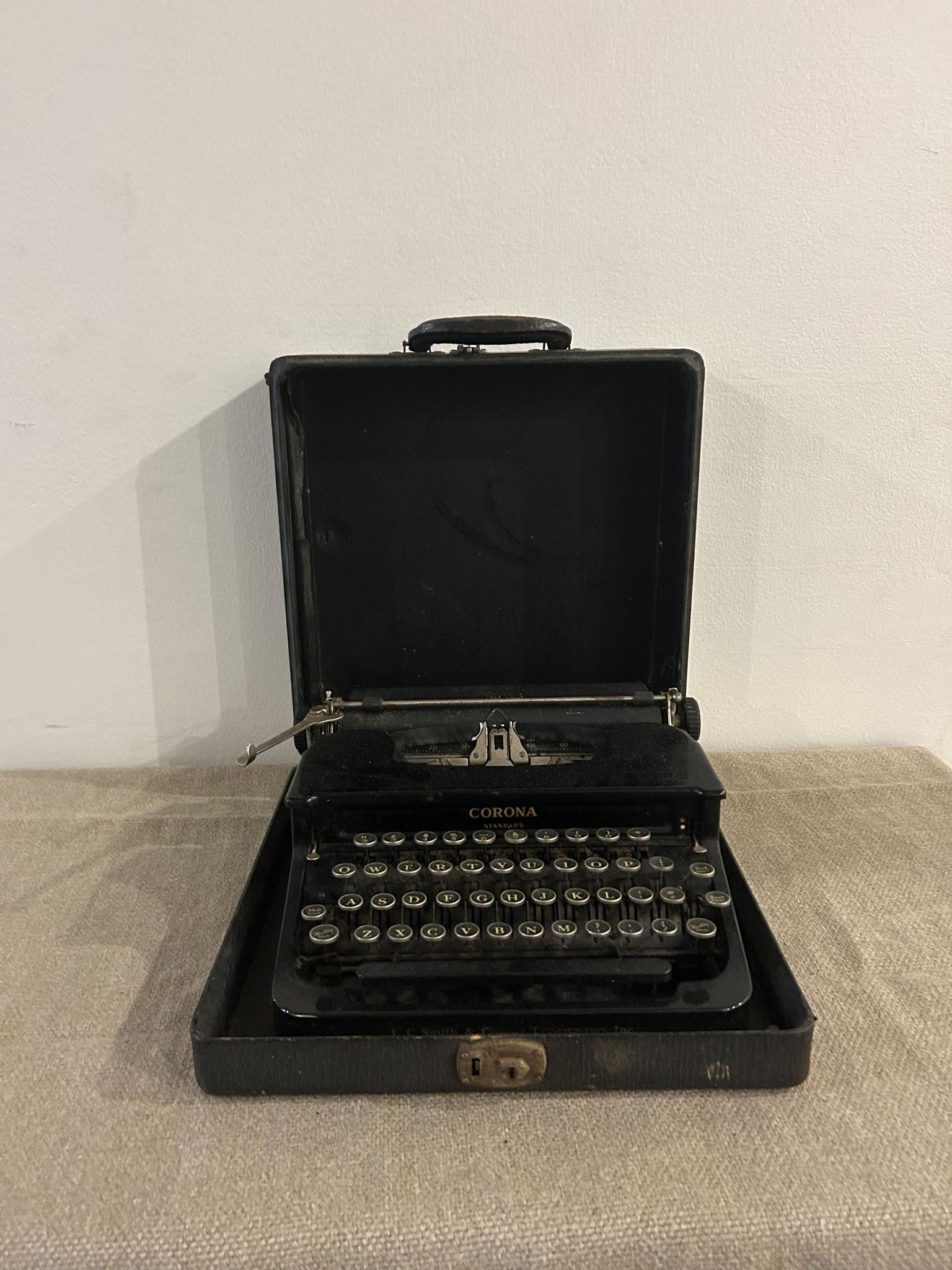 The Black Typewriter is a vintage portable model displayed open in its carrying case on a beige surface, with keys and handle visible against a plain white wall.