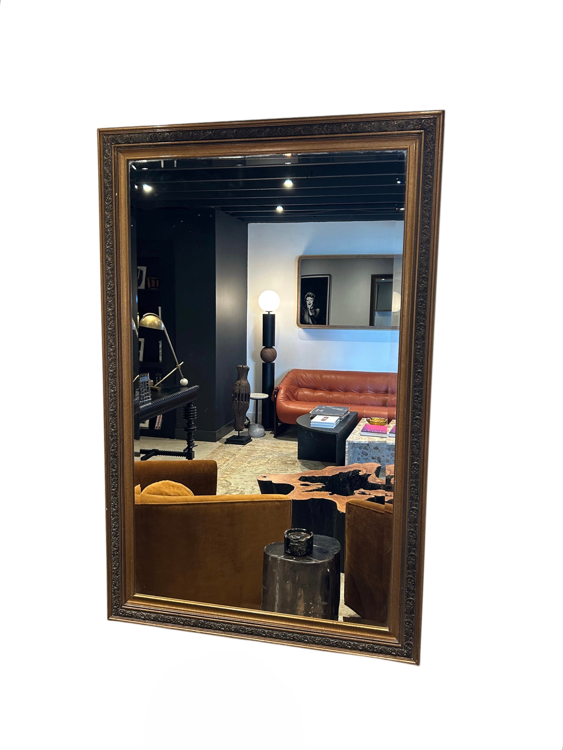 The Gilt Frame Leaner Mirror (43.5"w x 67.5"h) reflects a chic living room with a brown leather sofa, armchairs, a lamp, and framed art. Its ornate rectangular design stands out against the mostly white background.
