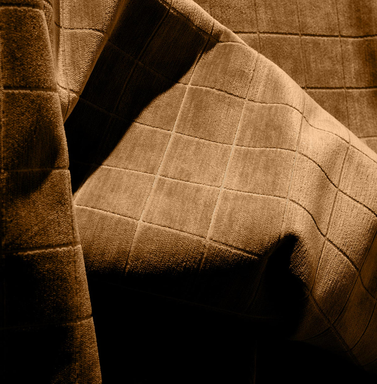 Close-up of the Cumin Grid Pillow, featuring brown fabric with a subtle grid pattern, draped over a feather pillow and softly lit to showcase its texture and elegant folds.