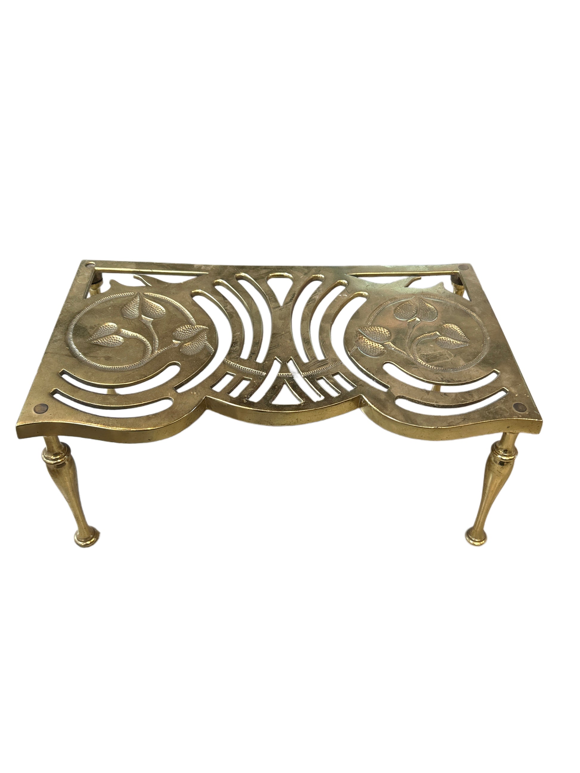 The Brass Lift is a vintage brass trivet featuring ornate cut-out floral and curved patterns, supported by four slender legs.