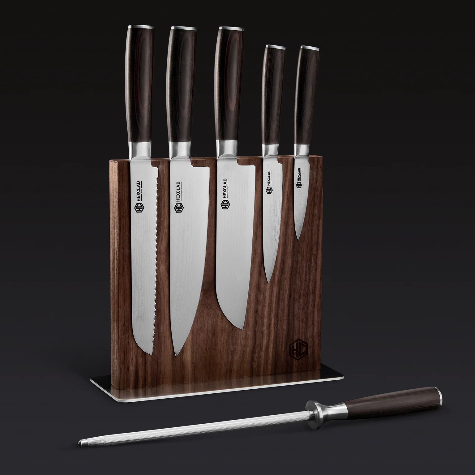 The Hexclad Steel Knife Block features five kitchen knives with dark handles displayed upright in a wooden magnetic block, plus a metal honing rod in front, all set against a dark background.