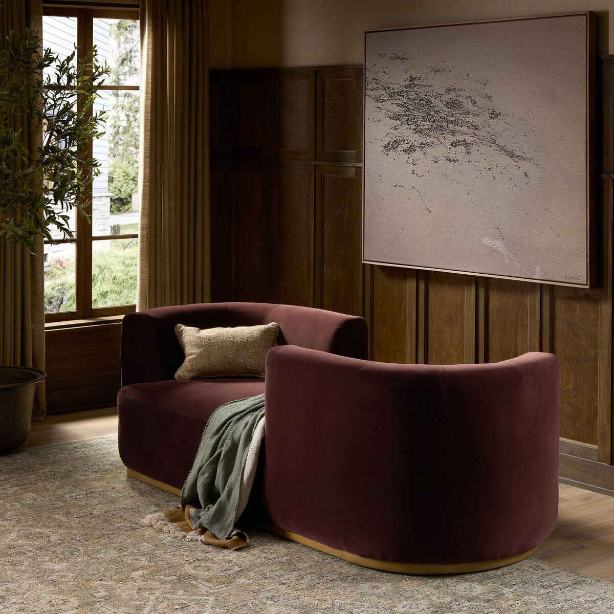 The Deandra Tete A Tete Chaise, a stylish dual-sided maroon velvet 90.50" sofa, sits in a cozy room with dark wood paneling, a large window, potted plant, neutral rug, abstract art, plus a pillow and blanket on the chaise.