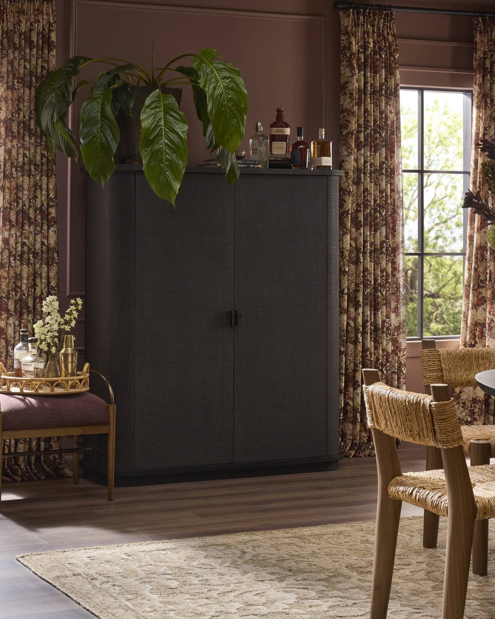 A cozy room with the Berlin Cabinet (55.00"w x 20.00"d x 62.00"h) topped with bottles and a leafy plant, floral curtains by tall windows, a bar cart with glassware, cushioned bench, wooden dining chairs, and a beige rug on wood floors.