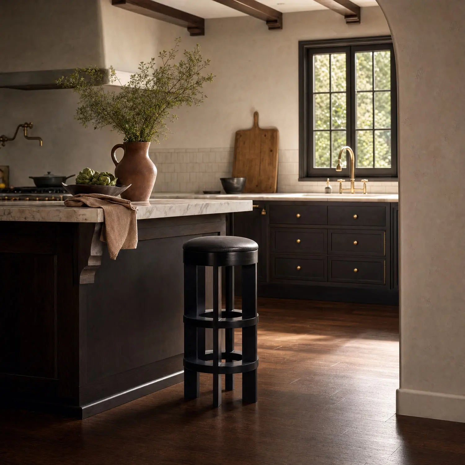 Sunlight streams into a cozy kitchen with dark wood cabinets, a marble island, and a Swivel Bar Stool featuring a hand-rubbed black finish and leather seat. A brass faucet sits below the window, lighting up the hardwood floor.
