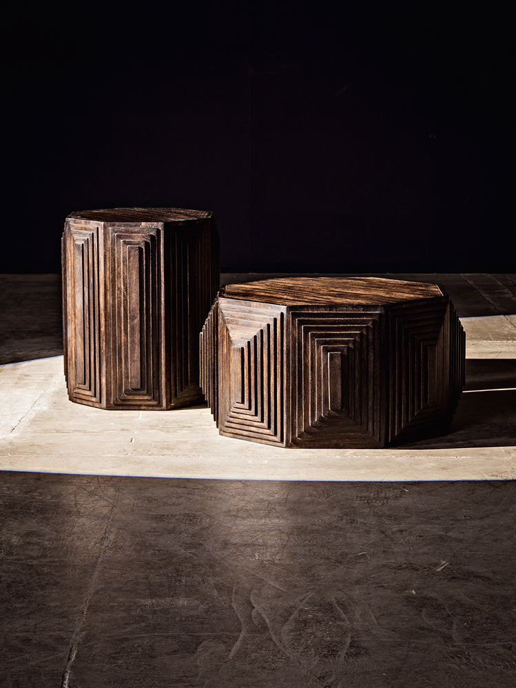 Two Moore Side Tables in Ebony, handcrafted from walnut with angular carved patterns, are displayed on a textured concrete floor and illuminated by focused light against a dark background.