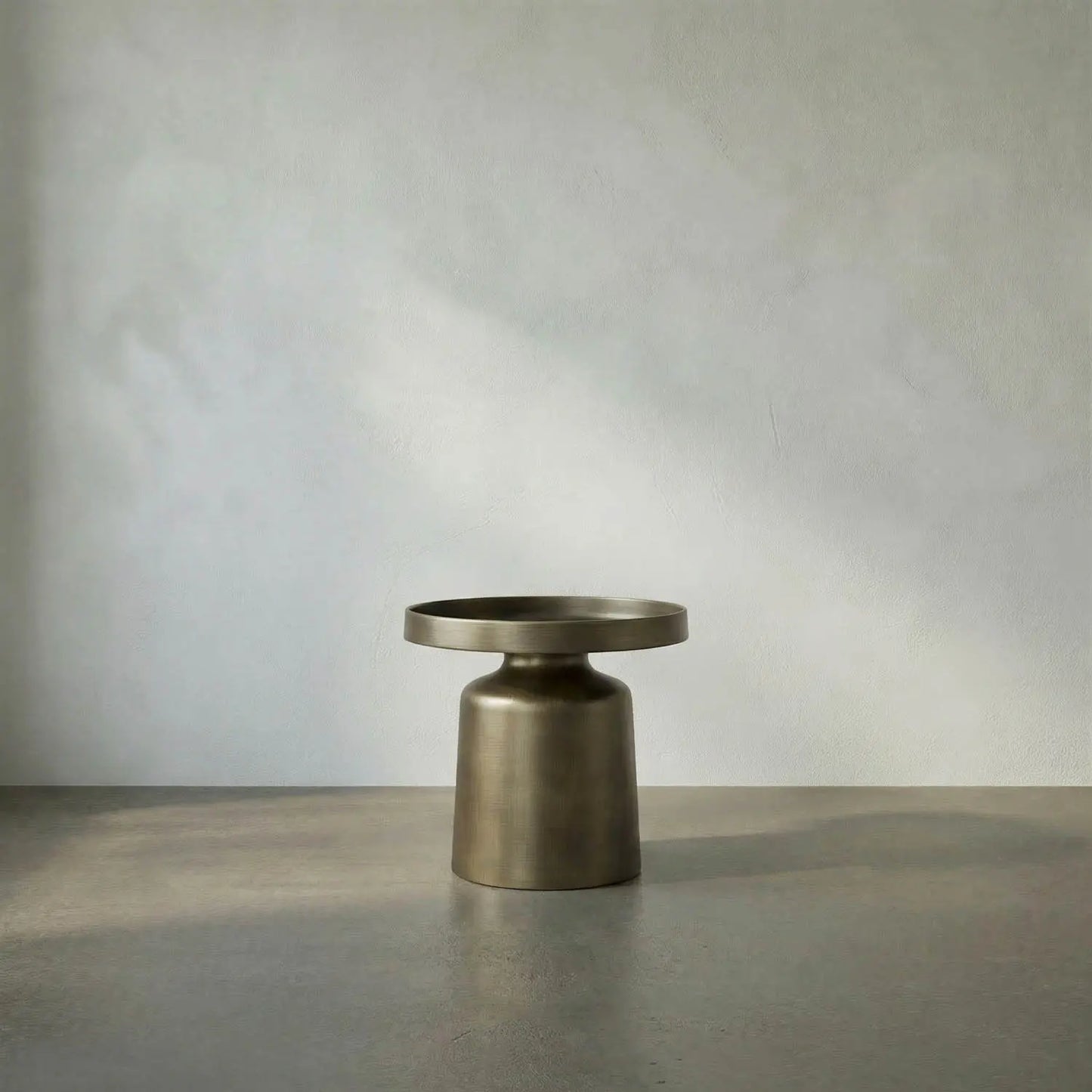 The Aged Brass Tray Top Side Table is a small, round metallic piece featuring a wide, flat top and a cylindrical base, positioned on a gray surface against a plain light-colored wall. Measuring 19W x 19D x 18.5H, it offers an elegant yet practical addition to any modern interior.