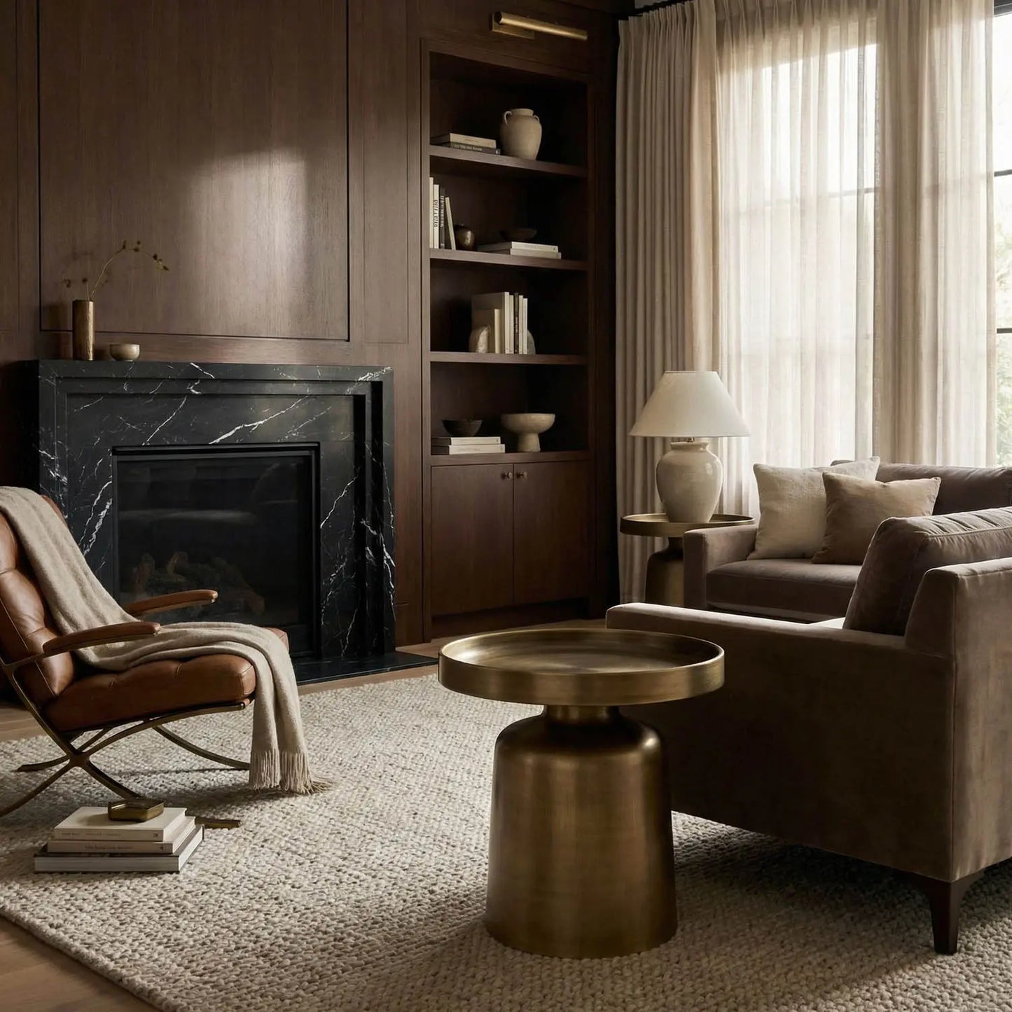 A cozy living room with dark wood walls, a black marble fireplace, built-in shelves displaying books and vases, a gray sofa, a leather chair, and the Aged Brass Tray Top Side Table (19W x 19D x 18.5H). The space is finished with a lamp and tall beige curtains, all thoughtfully arranged to create an inviting atmosphere.