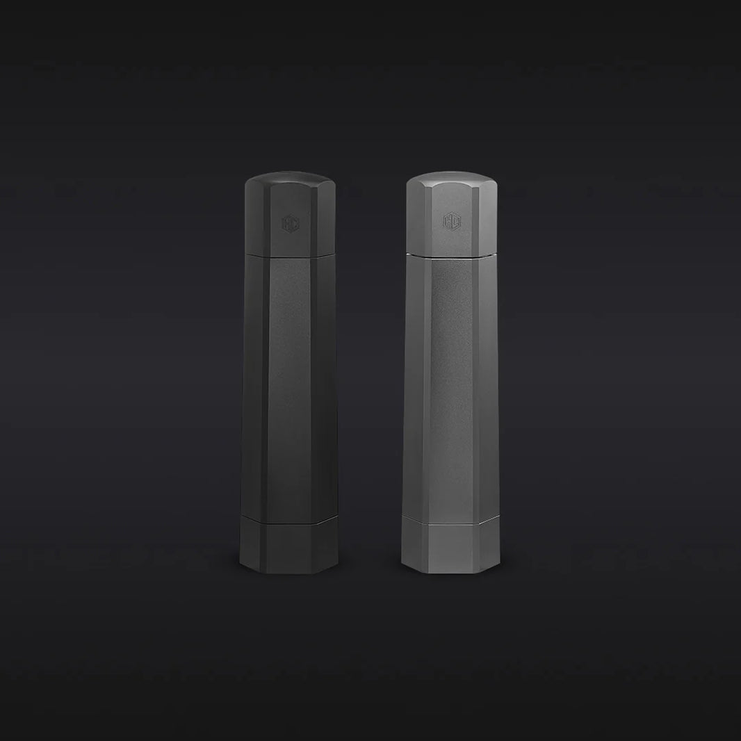 The Hexclad Salt and Pepper Grinder Set features two tall, hexagonal containers in black and gray with subtle embossed logos near the top, displayed side by side against a dark gradient background.