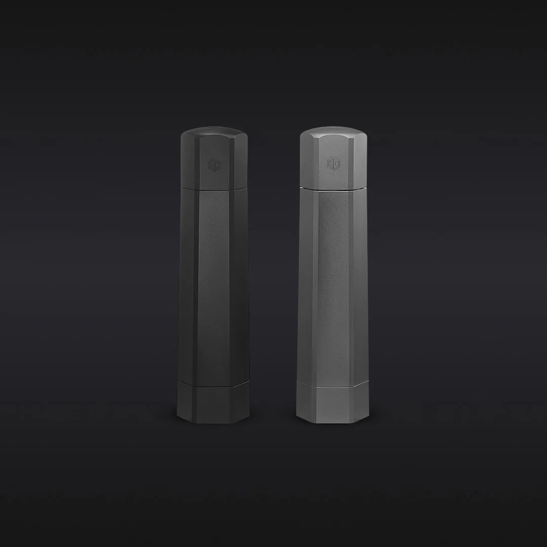 The Hexclad Salt and Pepper Grinder Set features two tall, hexagonal containers in black and gray with subtle embossed logos near the top, displayed side by side against a dark gradient background.