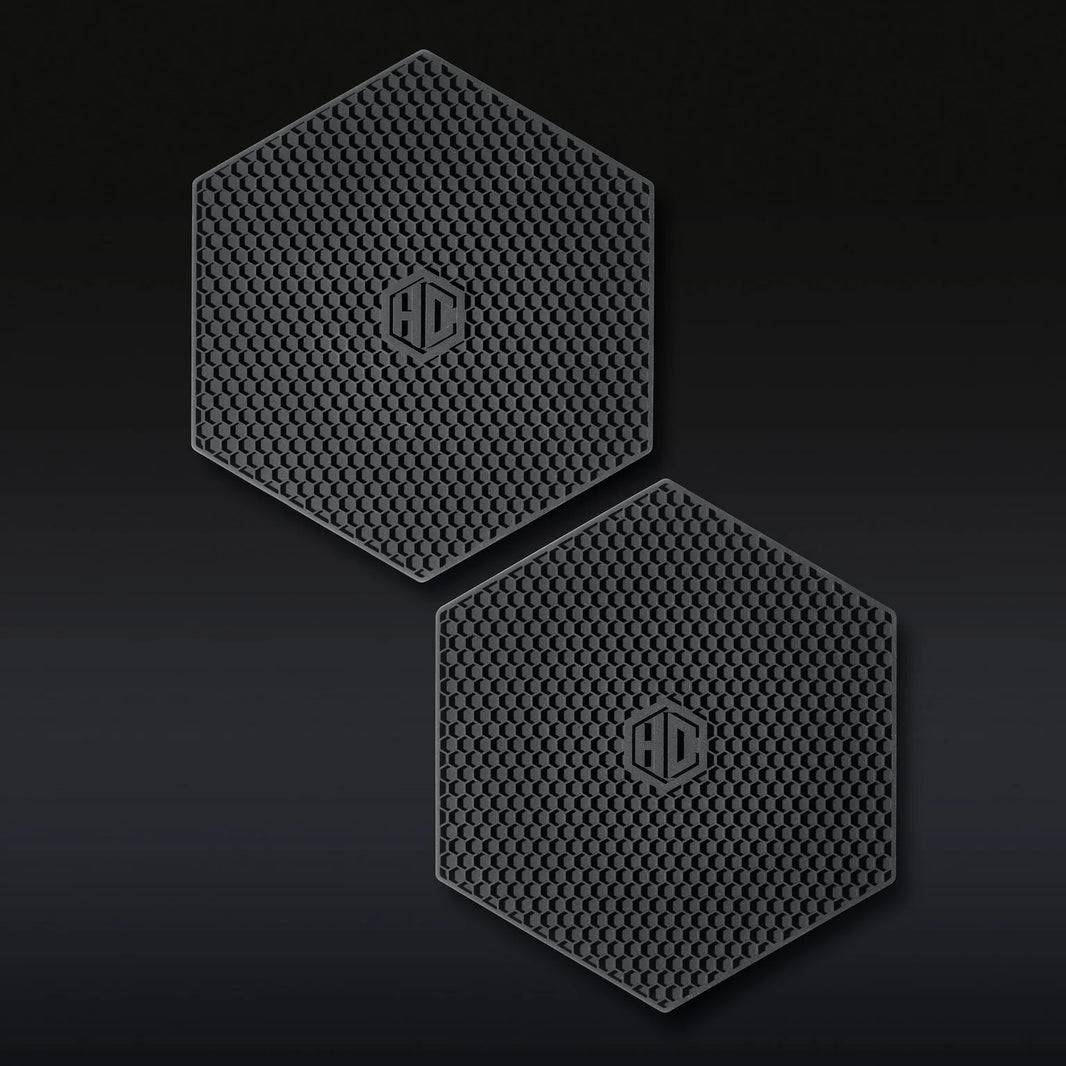 The Hexclad Trivet Set of 2 features black, hexagonal silicone pads with a honeycomb texture and a raised HD logo in the center, shown side by side on a dark background.
