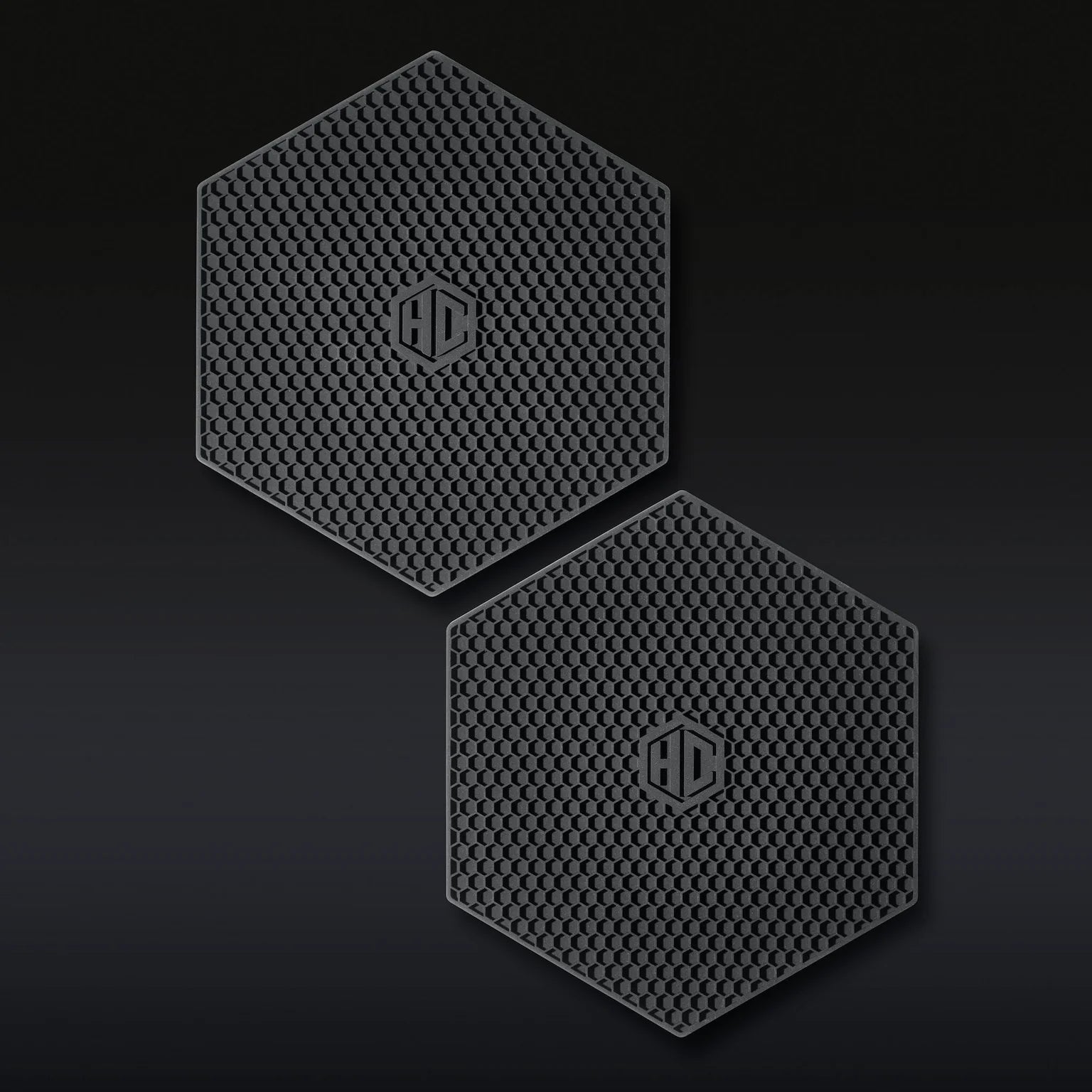The Hexclad Trivet Set of 2 features black, hexagonal silicone pads with a honeycomb texture and a raised HD logo in the center, shown side by side on a dark background.
