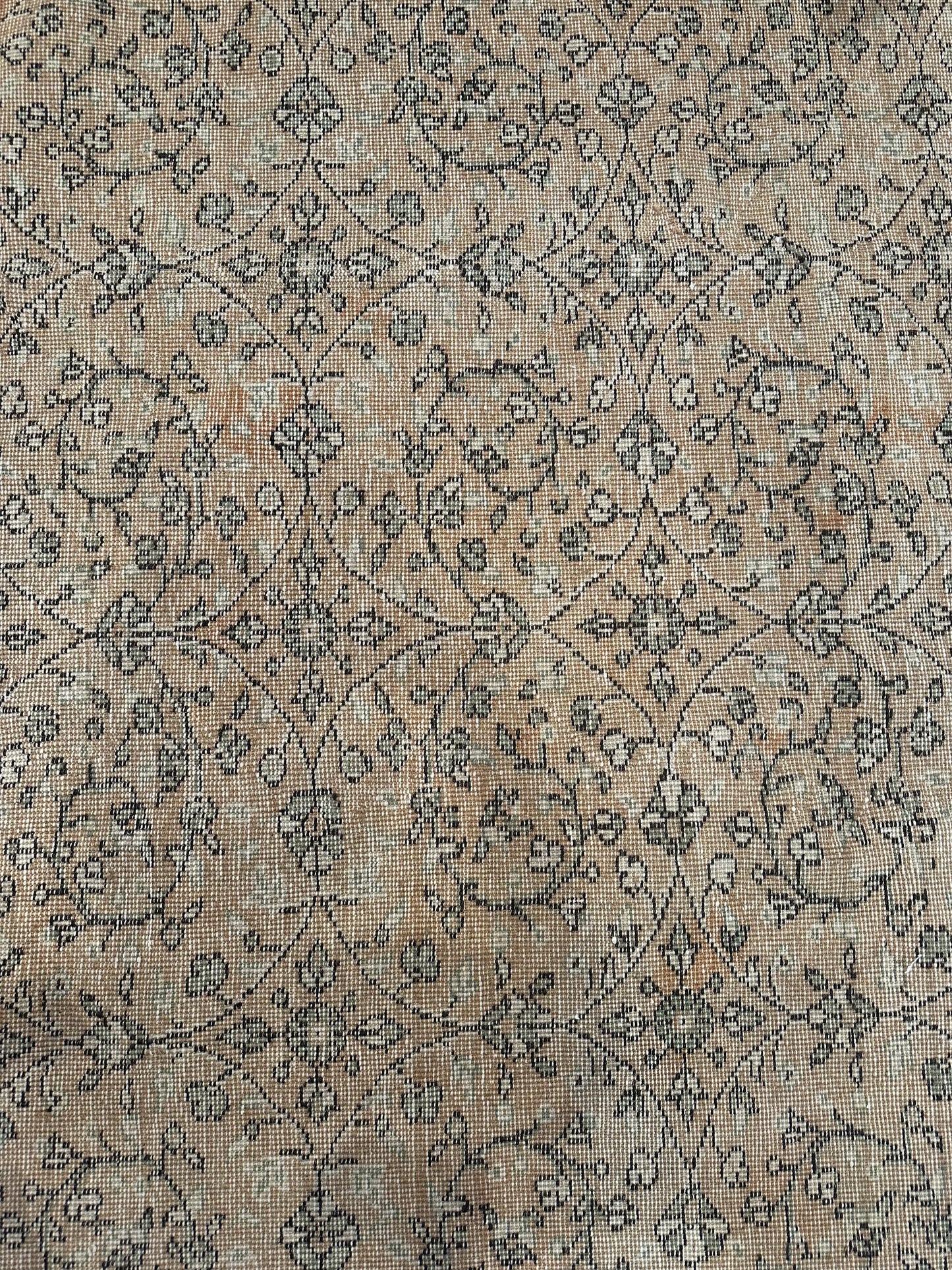 A close-up of the Reversible Vintage Rug reveals a faded beige background with intricate, repeating floral and vine motifs in muted brown and gray, showcasing its fabric’s texture and charming vintage wear.
