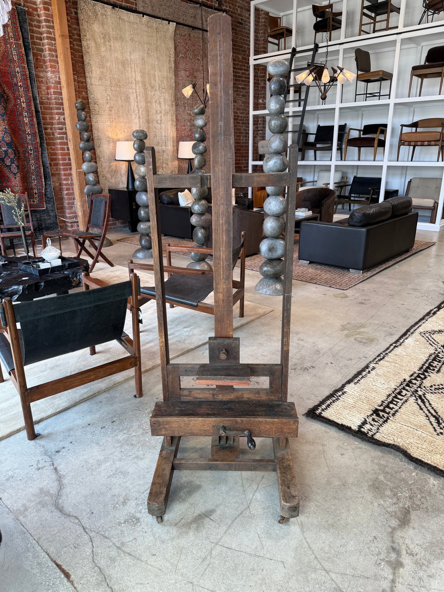 The 1800s Wood Art Easel, featuring a vertical post and adjustable supports on wheels, stands in a modern store surrounded by furniture, rugs, and chairs.