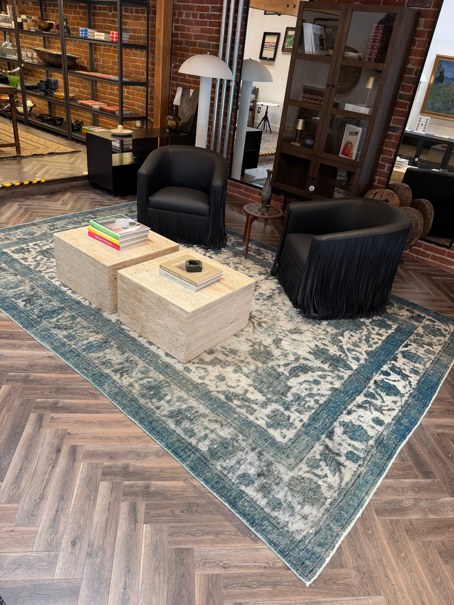 A Vintage Blue Distressed Wool Area Rug with a blue and cream distressed pattern and traditional ornate border, centered under two black fringed chairs and two square stone coffee tables in an 8x12 garden shed.