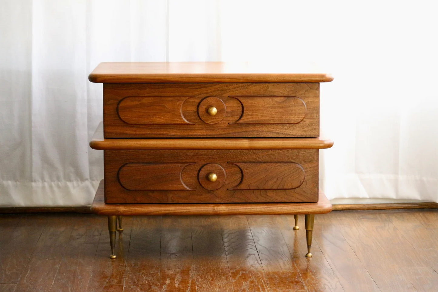 The Ronson Nightstand features solid walnut construction, dovetail drawer boxes, rounded edges, and patinated brass accents on knobs and tapered legs, shown on a wooden floor in front of white curtains.