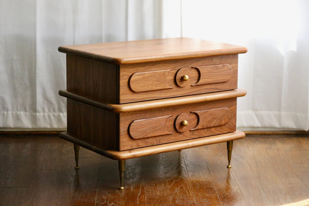 The Ronson Nightstand, crafted from solid walnut, features two dovetail drawers with circular brass knobs and wood detailing. It stands on tapered patinated brass accents, set on a wooden floor before white curtains.
