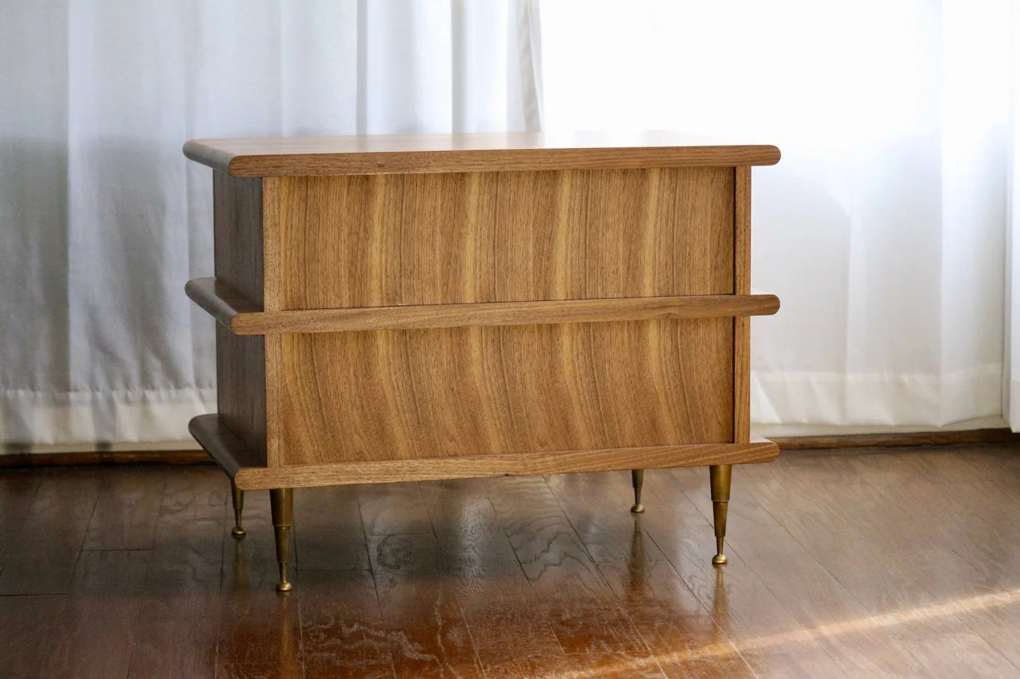 The Ronson Nightstand, a mid-century modern piece with clean lines, solid walnut construction, patinated brass accents, and dovetail drawer boxes, sits on a wooden floor in front of white curtains.