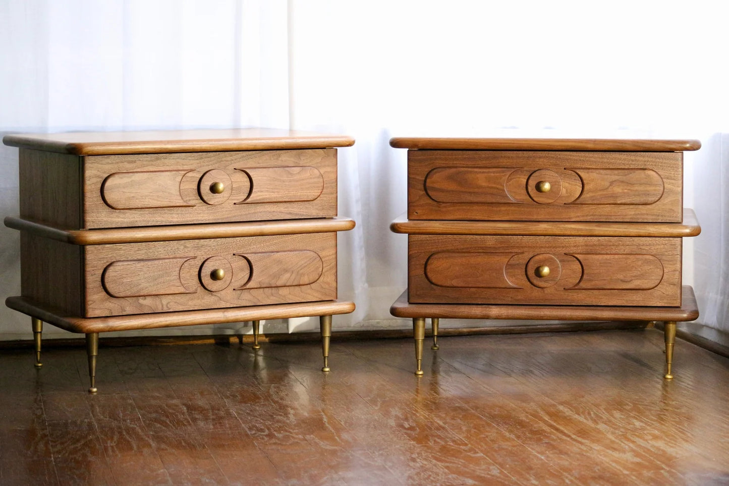 A pair of Ronson Nightstands in solid walnut with patinated brass legs, dovetail drawer boxes featuring circular patterns, and round brass knobs sit on a wooden floor beside a white curtain.