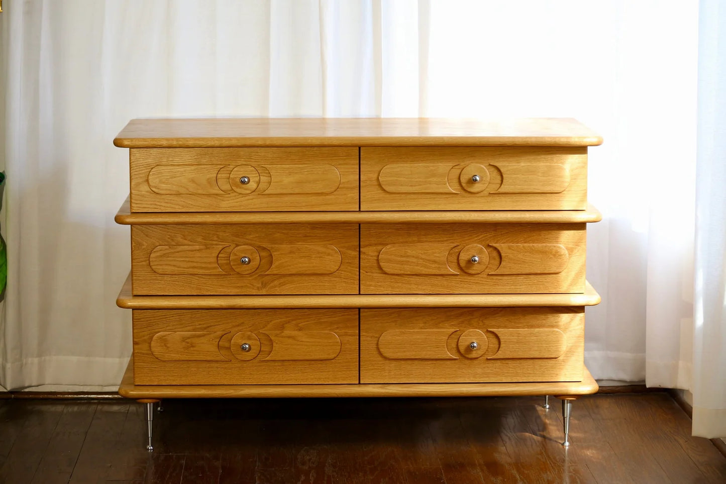 The Ronson Dresser, crafted from white oak with six dovetail drawers and circular handles, stands on metal legs against white curtains and a wooden floor.