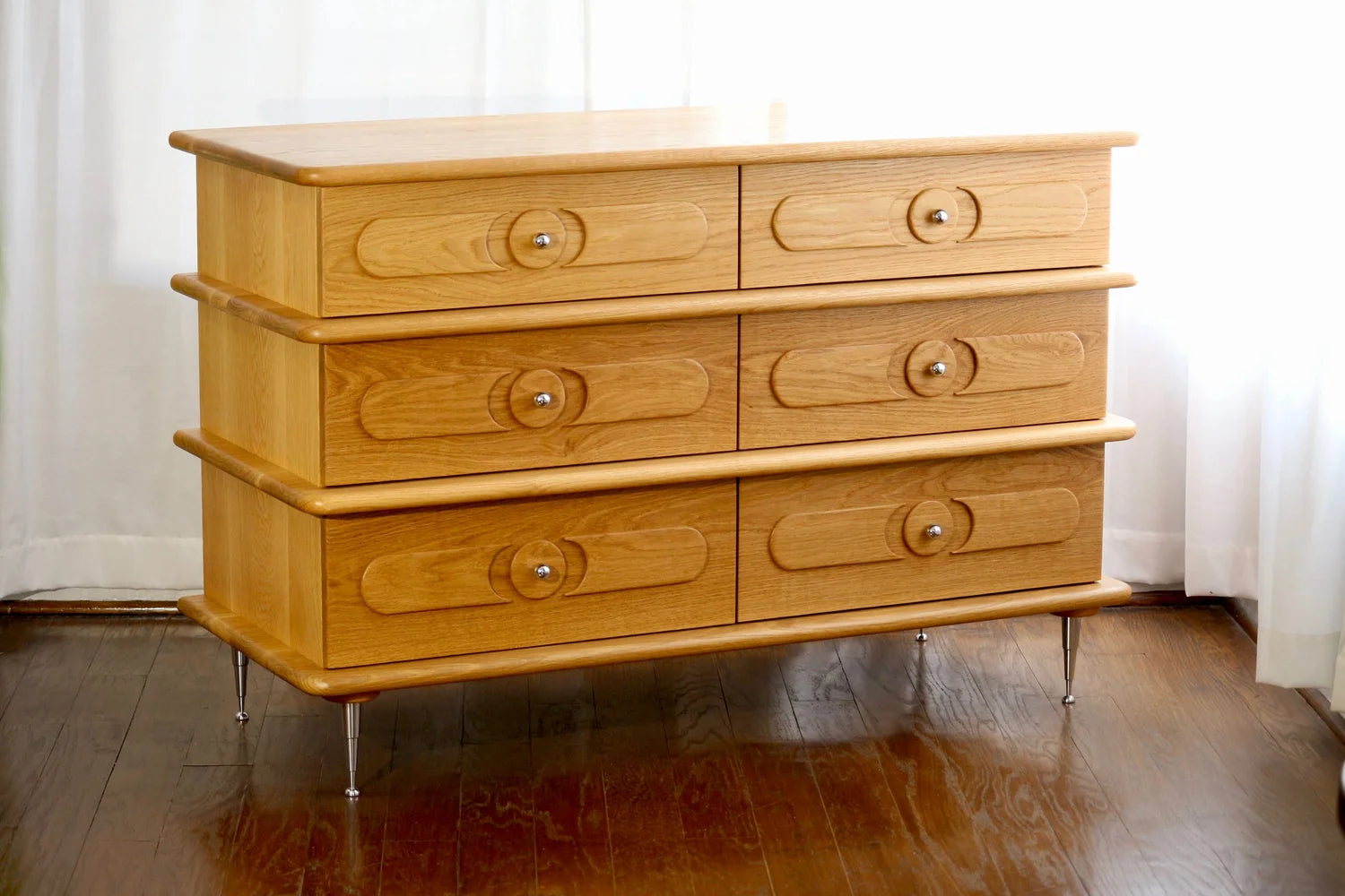 The Ronson Dresser, crafted from white oak, features six soft-close drawers with circular knobs and rounded edges. Standing on short metal legs, it sits on a hardwood floor near white curtains as sunlight filters gently in.