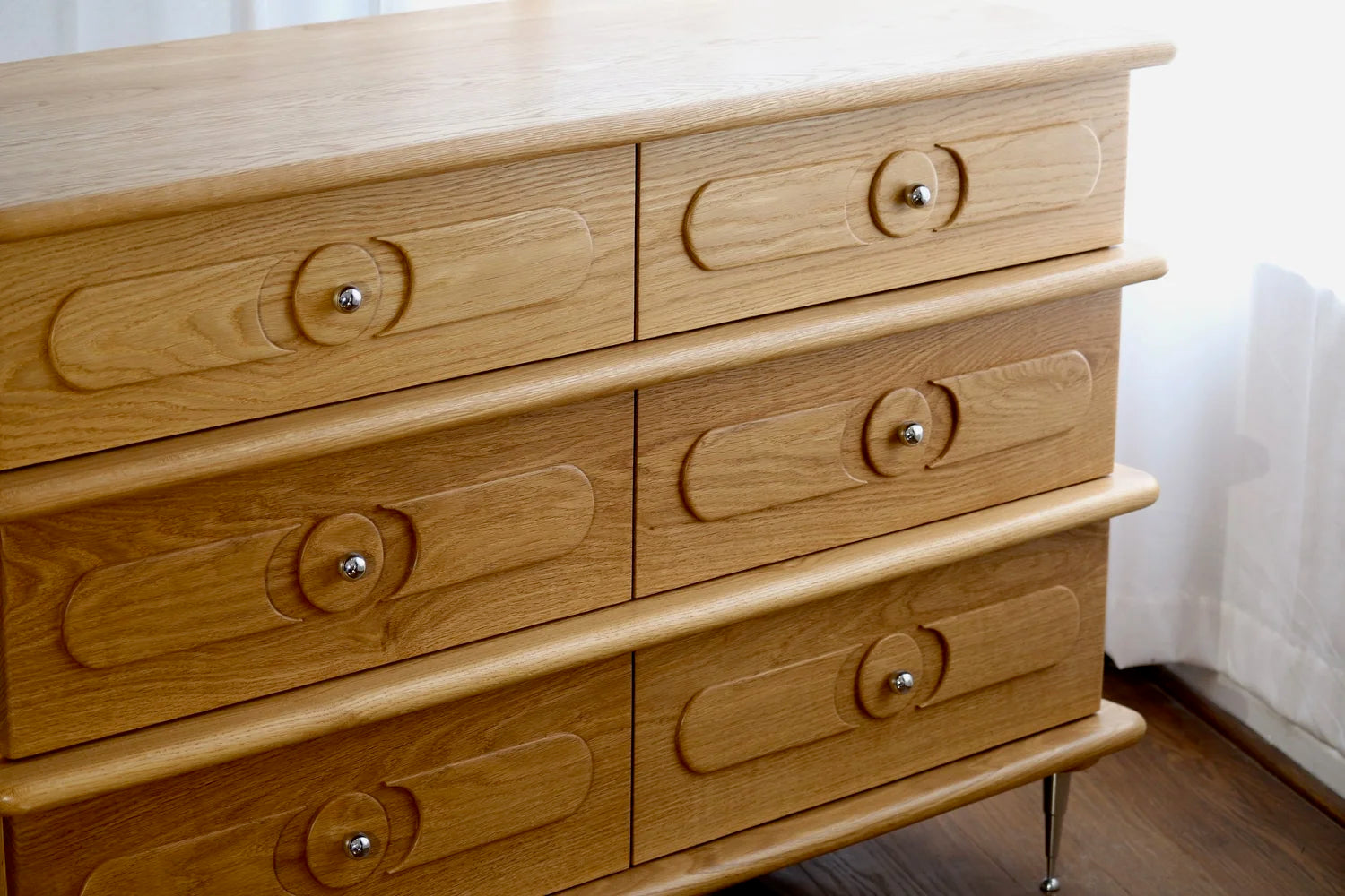 The Ronson Dresser, crafted from white oak, features six soft-close drawers with circular knobs on oval designs and stands on short metal legs beside a window with sheer white curtains.