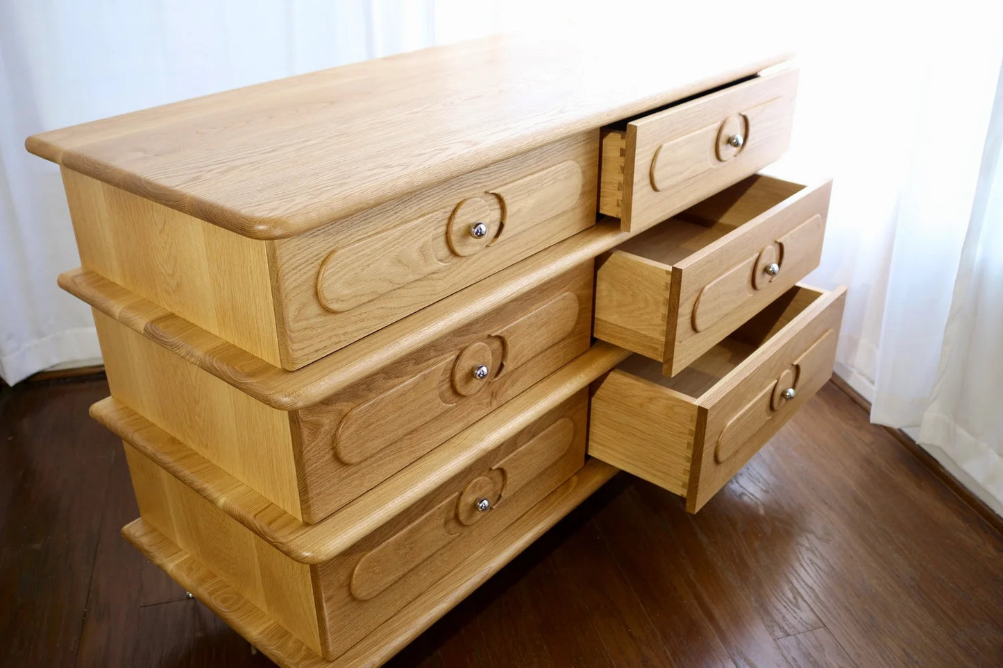 The Ronson Dresser, made of light white oak, features six drawers with round handles and dovetail construction. Three drawers are partially open. It stands on a wooden floor beside white curtains.