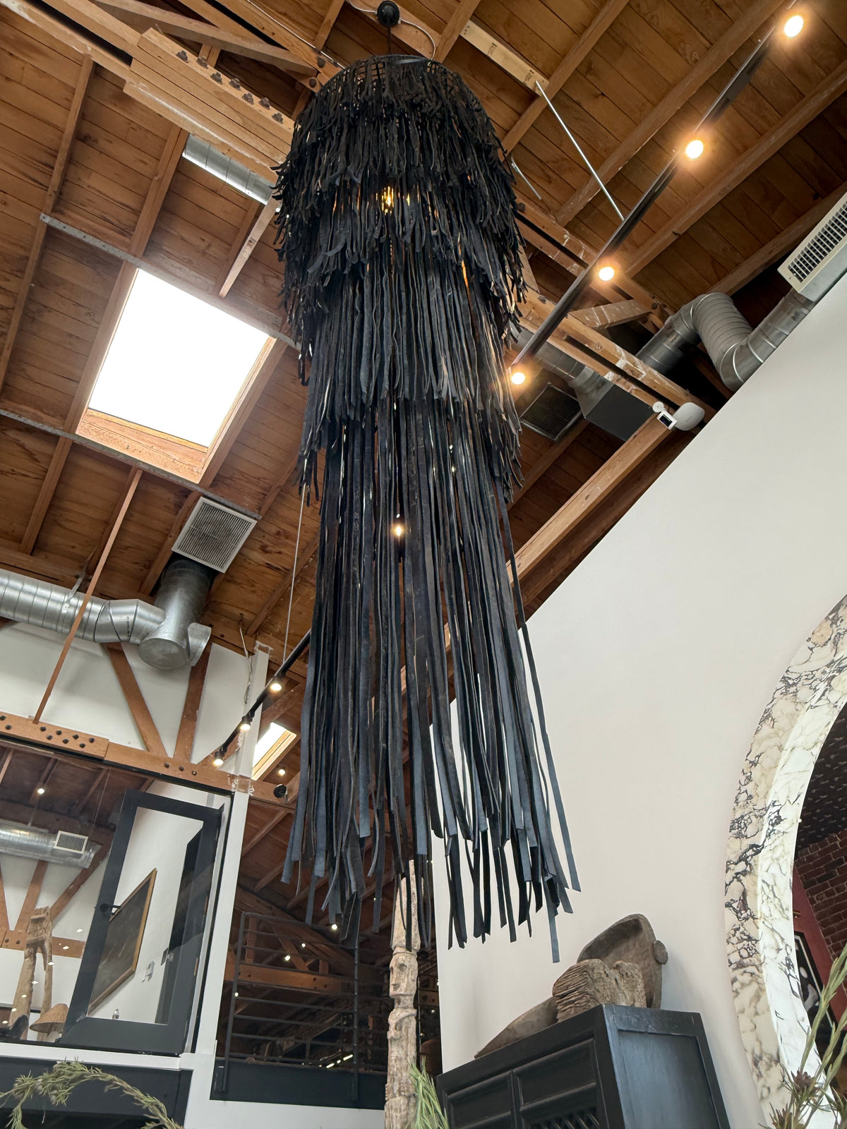 The Black Vegan Leather Fringe Chandelier hangs from a high wooden ceiling in an industrial-style space with exposed beams and ducts, its dramatic fringe shade creating a striking focal point.