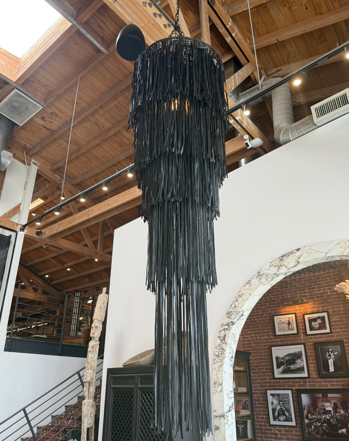 The Black Vegan Leather Fringe Chandelier hangs from a high, exposed wooden ceiling in a modern industrial space with brick walls, framed photos, and a marble archway.