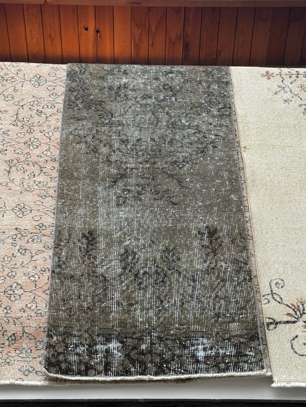 A Vintage Black Turkish Runner Rug with a textured pattern is displayed on top of two larger, lighter rugs with subtle designs, all arranged on a wooden floor to showcase their distinct sizes and dimensions.