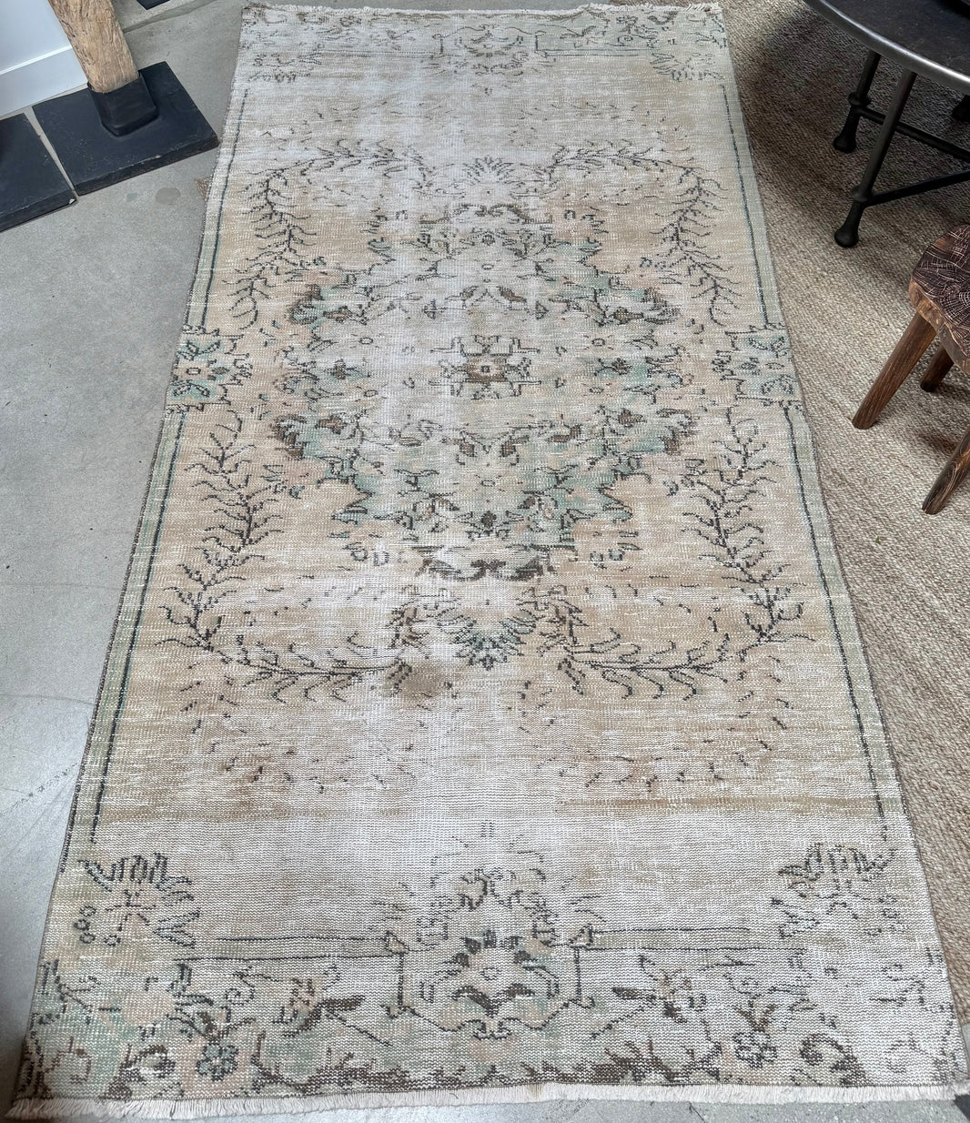 The Vintage Floral Motif Rug, measuring 9'6" x 4'8", displays faded beige, light blue, and green floral patterns. With its weathered, distressed appearance, this vintage piece is positioned on a concrete floor beside a woven mat, a chair, and a fireplace.