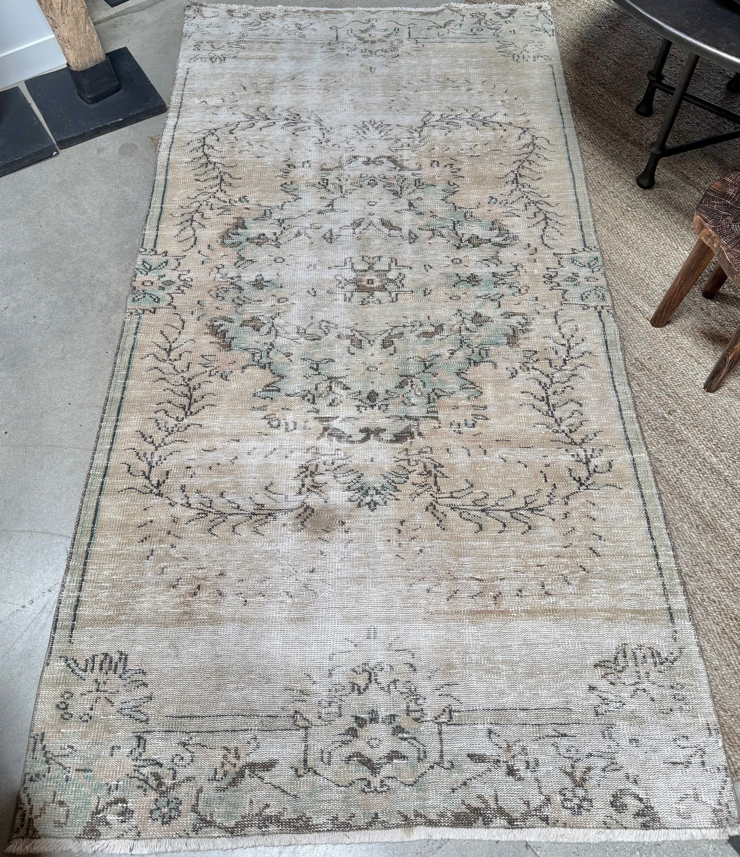 The Vintage Floral Motif Rug, measuring 9'6" x 4'8", displays faded beige, light blue, and green floral patterns. With its weathered, distressed appearance, this vintage piece is positioned on a concrete floor beside a woven mat, a chair, and a fireplace.