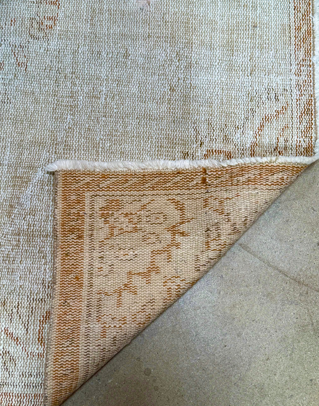A corner of the Vintage Cream/Sienna Turkish Rug is flipped over, revealing a brown and cream patterned underside. Measuring 8'10" x 4'8", the rug rests on a smooth, light-colored floor.
