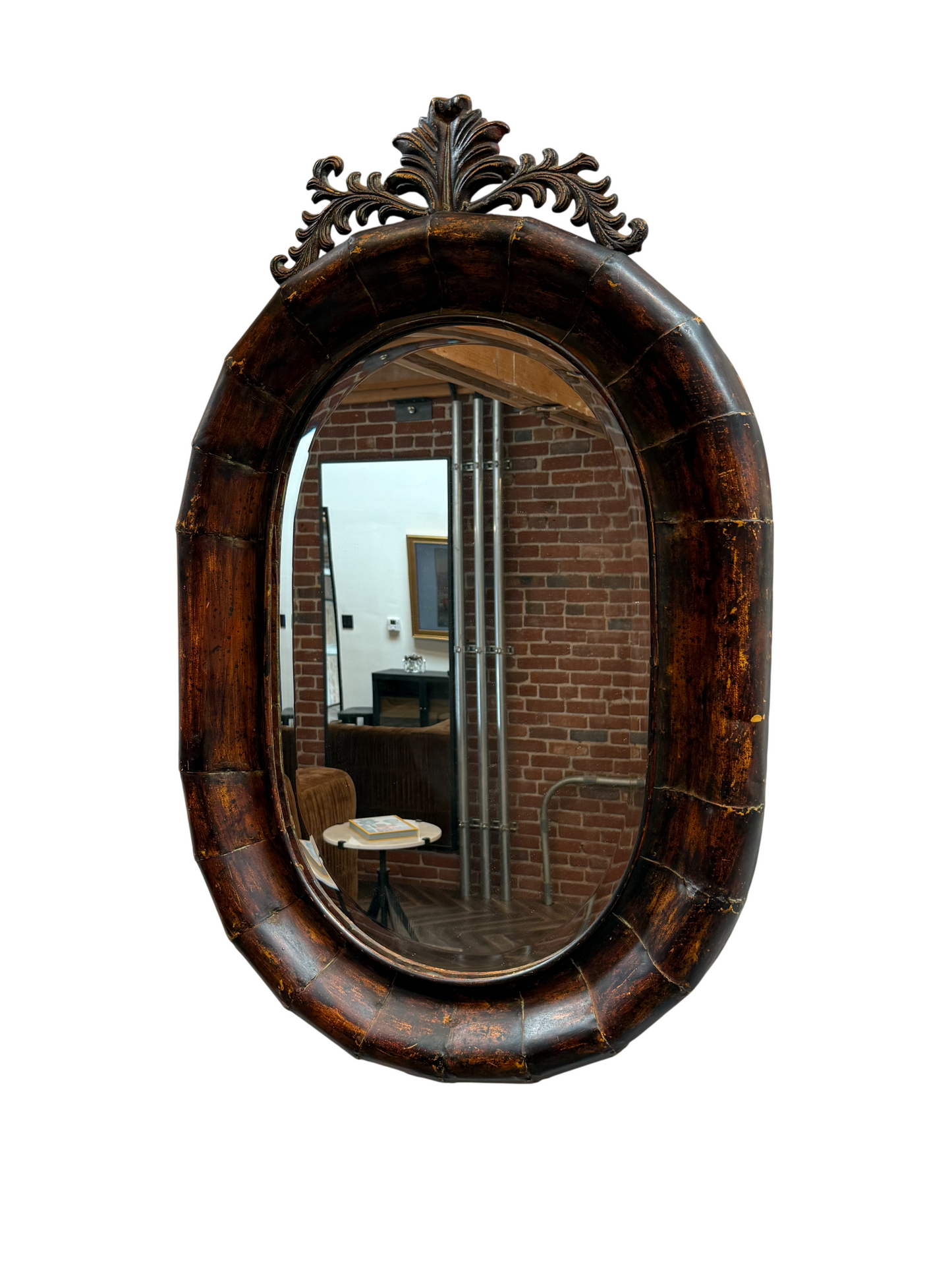The Vintage Crested Oval Mirror, measuring 24″ x 38″, features an ornate wooden frame and reflects a room with brick walls, a chair, metal pipes, and a round table with books, all set against a white background.